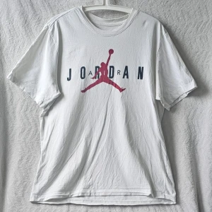 Vit Jordan t-shirt med logga - Säljer en vit t-shirt från Jordan med den klassiska Jumpman-loggan i rött och svart text på bröstet. T-shirten har korta ärmar och rund halsringning. Tillverkad i mjuk bomull för skön känsla och avslappnad passform.