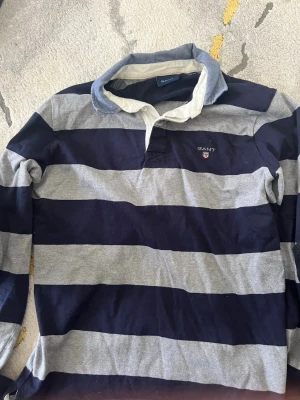 Randig rugbytröja från GANT - Säljer en klassisk långärmad rugbytröja från GANT i marinblått och grått med breda ränder. Skjortan har vit krage, knappar i halsen och GANT-logga broderad på bröstet. Perfekt för en avslappnad men ändå stilren look.