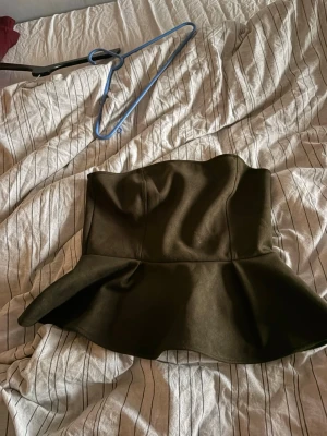Mörkgrön peplum tubtopp - Snygg mörkgrön tubtopp med peplumvolang nertill. Toppen är axelbandslös och har en figurnära passform upptill. Materialet känns mjukt och har en lätt struktur. Perfekt för att skapa en trendig och stilren look.