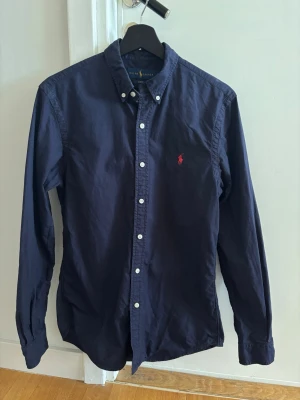 Mörkblå skjorta från Ralph Lauren i nyskick, S - Klassisk mörkblå skjorta i slim fit-modell från Ralph Lauren med button-down krage och vit knappstängning. Använd två gånger. Drar mer åt XS än S. Mått: 📏 bakstyckets längd från nacke till plaggets nederkant = 78 cm, ärmlängd (från mitten av nacken till nederkant ärm) = 87 cm, axelbredd = 42 cm, bröstmått = 96 cm, hals = 37 cm 