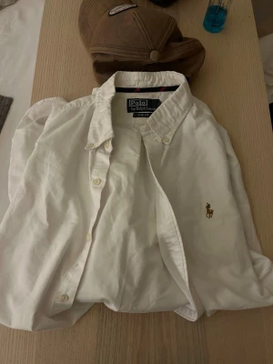 Vit skjorta från Polo Ralph Lauren - Klassisk vit skjorta från Polo Ralph Lauren med broderad logga på bröstet. Skjortan har knappar framtill och button-down krage. Tillverkad i bomull och har lång ärm. Perfekt till en clean och stilren look. Pris kan diskuteras privat m