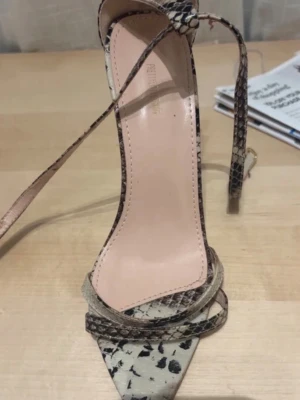 Ormmönstrade stilettklackar med remmar - Säljer ett par snygga pumps med ormmönster i beige och svart. Skorna har tunna remmar som går runt ankeln och en spetsig tå. Höga stilettklackar ger en elegant look. Materialet ser ut att vara konstläder med präglad struktur.