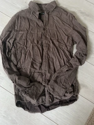 Brun skjorta från Dressmann - Brun långärmad skjorta från Dressmann i mjukt bomullstyg. Klassisk krage och knäppning framtill. Perfekt för en avslappnad men ändå stilren look.
