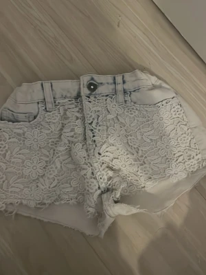 Coola Vita/ljusblå spetsdetaljerade jeansshorts - Säljer ett par vita jeansshorts med blommig spets framtill och råa kanter. Shortsen har fickor både fram och bak samt bälteshällor. Perfekta för sommaren och ger en söt och trendig look.