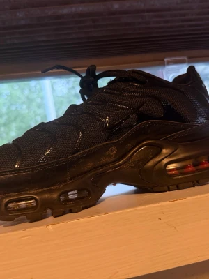 Nike Air Max Plus helsvart sneaker - Nike Air Max Plus i helsvart med synliga luftbubblor i sulan och vågformade detaljer på ovandelen. Skon har en chunky sula och är tillverkad i mesh och syntetmaterial. Snygg och sportig design med klassisk Nike-logga på sidan.