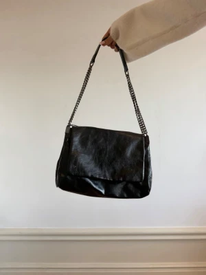 Svart axelväska med kedjerem - Stilren svart axelväska i glansigt skinnimitation med bred lockstängning och dubbla kedjeremmar. Väskan har en mjuk, något slouchy form och minimalistisk design utan synliga loggor. 