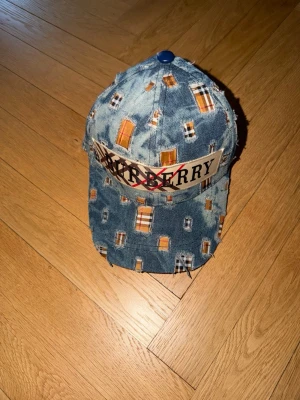 Burberry denimkeps med patchwork - Säljer en unik Burberry keps i blå denim med patchwork och slitna detaljer. Kepsen har ett coolt mönster med orange och vita rutor samt en stor Burberry-logga framtill. Justerbar rem bak i svart läder med präglad logotyp. Perfekt för dig som vill sticka ut!
