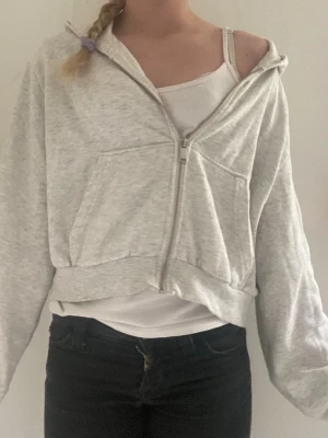 Ljusgrå zip hoodie med huva - En ljusgrå hoodie med dragkedja framtill och två stora fickor. Modellen är croppad och har en avslappnad passform. Tillverkad i mjukt sweatshirtmaterial och har en klassisk huva. Perfekt för en chill och enkel stil.