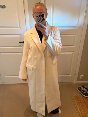 Lång vit dubbelknäppt kappa - Stilren lång vit kappa med dubbelknäppning och breda slag. Kappan har två fickor framtill och en rak, elegant passform. Perfekt för dig som vill ha en klassisk och trendig look. Från nakd, storlek 36