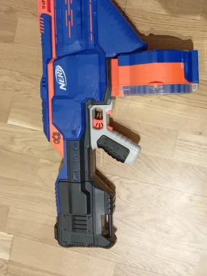 Nerf Elite Infinus Blaster - Säljer en Nerf Elite Infinus Blaster i blått, orange och svart plast. Den har ett stort trummagasin och ergonomiskt handtag. Perfekt för intensiva Nerf-strider och enkel att ladda om tack vare det smarta magasinet.