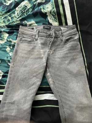 Grå jeans från Jack & Jones - Snygga gråa och svarta jeans från Jack & Jones med knäppning framtill och klassisk femficksdesign.  raka ben. Modellen är slim/glenn på bägge och midjan är 29 och längd 32. Båda för 700 aldrig använda