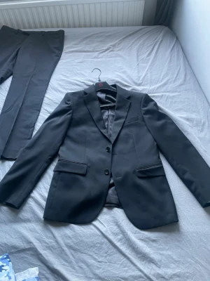 Svart klassisk kostym med kavaj och byxor - Säljer en svart kostym med klassisk kavaj och matchande byxor. Kavajen har två knappar, smala slag och fickor med lock. Kostymen är helfärgad och har en stilren, tidlös look som passar till många tillfällen.