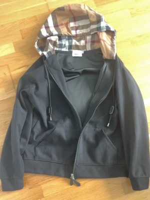 Svart hoodie från Burberry med dragkedja - Svart hoodie från Burberry med klassiskt rutigt foder i huvan och dragkedja framtill. Hoodien har fickor på magen, justerbara snören med metalländar och ribbade muddar. Perfekt för dig som gillar stilrena plagg med exklusiva detaljer.