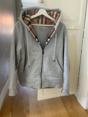 Grå hoodie från Burberry med dragkedja - Snygg grå hoodie från Burberry med klassiskt rutigt foder i huvan. Dragkedja framtill, två fickor och justerbar huva med snören. Perfekt för en chill och stilren look. Materialet är mjukt och bekvämt, troligen bomullsblandning.