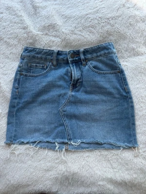 Blå jeanskjol  - Säljer en klassisk blå jeanskjol med fem fickor och rå, fransig kant nedtill. Kjolen har normal passform och stängs med knapp och dragkedja framtill. Perfekt till en avslappnad och trendig look.