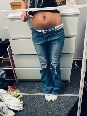 Ljusblå bootcut jeans med slitningar - Säljer ett par ljusblå bootcut jeans med slitna detaljer på knäna och klassisk femficksdesign. Jeansen har låg midja och är tillverkade i denim med lite stretch för skön passform. Perfekta för en avslappnad och trendig look.