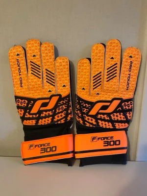 Pro Touch målvaktshandskar orange - Snygga målvaktshandskar från Pro Touch i orange och svart med mönstrad ovansida och breda kardborreband med texten Force 300. Handflatan är ljus och slät för bra grepp. Perfekta för fotbollsträning eller match.