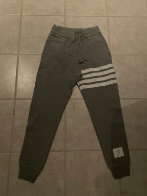 Gråa mjukisbyxor Thom Browne - Gråa mjukisbyxor från Thom Browne med ribbad struktur, vita ränder på ena benet och justerbar dragsko i midjan. Byxorna har muddar vid bensluten och en vit patch med logga på benet. Perfekta för en chill och stilren look. Storlek 0 (S)