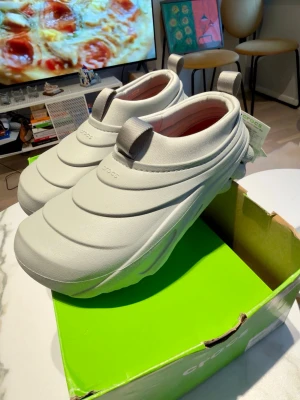Grå chunky tofflor från Crocs - Säljer ett par gråa Crocs tofflor med chunky, futuristisk design och räfflad sula. Tillverkade i lätt och slitstarkt syntetmaterial med dragflikar både fram och bak för enkel påtagning. Perfekta för dig som gillar en modern och bekväm stil.