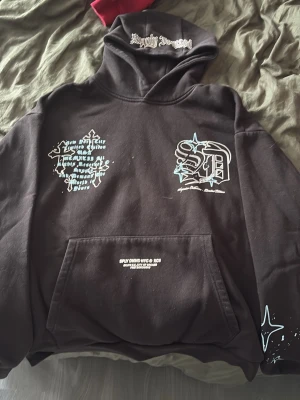 Svart hoodie Supply & Demand - Säljer en svart hoodie från Supply & Demand med tryck i vitt och ljusblått på bröstet och huvan. Hoodien har stor magficka och coola grafiska detaljer samt text på både framsidan och huvan. Perfekt för dig som gillar streetwear och unik design.
