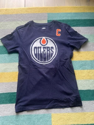 Edmonton Oilers McDavid t-shirt - Mörkblå t-shirt från Adidas med stort Edmonton Oilers-tryck på bröstet och McDavid 97 på ryggen. Klassisk sportig look med korta ärmar och rund hals. Perfekt för hockeyfans som vill visa sitt stöd för laget.