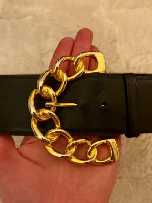 Coolt vintageskärp ESCADA - Escada 1980-tal – Svart läderbälte med guldkedja – Made in Western Germany Ett sällsynt och bärbart stycke lyxmodehistoria från 1980-talet. Detta äkta Escada-bälte härstammar från märkets tidiga år under Margaretha Ley. Storlek: 68-74 cm / XS. Material: Äkta läder Spänne: Överdimensionerat kedjelänkspänne i guldfärgad metall Färg: Svart / Guld Referens: 42/09320/900 Tillverkningsland: Västtyskland (före 1990) Skick: Gott vintage-skick.