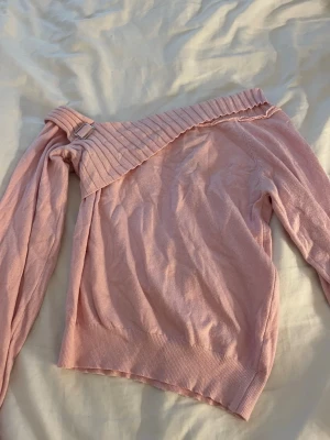 Ljusrosa off shoulder stickad tröja - Trendig ljusrosa stickad tröja med off shoulder-design och bred ribbad kant upptill. Tröjan har en dekorativ metallspänne på ena axeln och långa ärmar. Perfekt för dig som vill ha en unik och snygg stickad topp.