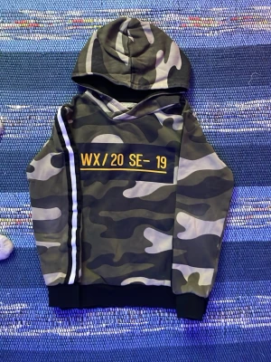 Camo hoodie med text och rand - Snygg hoodie i camomönster med huva och svartvita ränder längs ena ärmen. På bröstet står det 'WX/20 SE-19' i gul text. Muddar vid ärmslut och nederkant. Perfekt för dig som gillar streetwear och vill sticka ut lite extra.