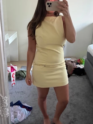 Ljusgul kort klänning med bälte - Snygg ljusgul kort klänning med minimalistisk design och ett smalt bälte i midjan. Klänningen är ärmlös och har en rak, figurnära passform som ger en stilren look. Perfekt för sommarens alla tillfällen.