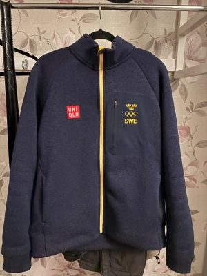 Blå Uniqlo OS-jacka med gul dragkedja - Mörkblå jacka från Uniqlo med gul dragkedja, OS-märke och SWE-tryck på bröstet samt svensk flagga på ärmen. Jackan har hög krage, bröstficka med dragkedja och sportig look. Perfekt för dig som gillar sport och stilren design. En del av Milano Cortina 2026 kollektionen, priset går att diskutera!