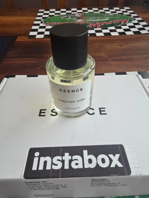 ESSNCE Forever June Eau de Parfum - ESSNCE Forever June Eau de Parfum är en stilren parfym i en rund, genomskinlig glasflaska med svart lock. Doften har inslag av citrus, ros och träiga toner. Flaskan rymmer 50 ml och har en minimalistisk vit etikett med svart text.