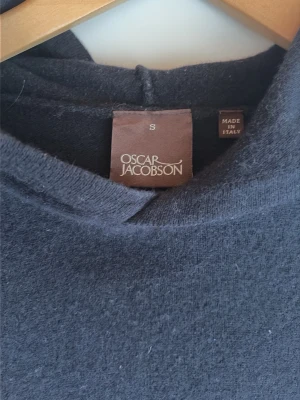 Mörkblå stickad hoodie Oscar Jacobson - Mörkblå stickad hoodie från Oscar Jacobson, tillverkad i Italien. Tröjan har lång ärm, ribbstickade muddar och en luva. Perfekt för dig som vill ha en stilren och bekväm look. Materialet är mjukt och ger en skön känsla.