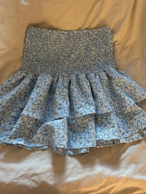 Blommig blå volangkjol - Supersöt kort blå kjol med volanger och blommigt mönster i vitt och grönt. Kjolen har smock i midjan för en bekväm passform och är perfekt för vår och sommar. Materialet känns lätt och luftigt.