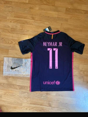 Barcelona Neymar Jr matchtröja Nike - Snygg Barcelona matchtröja 2016/17 från Nike med Neymar Jr och nummer 11 på ryggen. Tröjan är mörkblå med rosa detaljer, har officiella klubbmärket, Qatar Airways-tryck och Champions League-märke på ärmen. Perfekt för dig som älskar fotboll och Barca. 