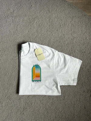 Vit Casa Blanca t-shirt med tryck - Vit t-shirt från Casa Blanca med färgglatt tryck framtill som föreställer en solnedgång i ett fönster. Klassisk passform och rund hals. Tillverkad i mjuk bomull och har korta ärmar. Perfekt för dig som gillar stilrena plagg med en twist. Den är också perfekt för sommaren. Fraktar så fort du har köpt📦🚚Aktiv 24/7✅ Hör av dig om du undrar något⁉️