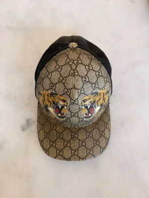 ÄKTA! Gucci keps med tigertryck - ÄKTA! Säljer en snygg Gucci keps med klassiskt GG-mönster i beige och brunt, två tigrar framtill och svart mesh baktill. 