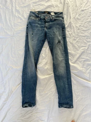 Slim fit jeans från Jack & Jones - Slim fit jeans från Jack & Jones i modellen Glenn. Jeansen är blå med slitningar och hål på benen för en trendig look. Klassisk femficksmodell med dragkedja och knapp. Perfekta för dig som gillar en smal passform och streetstil.