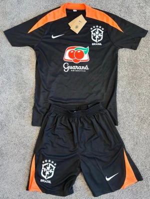Svart Brasilien träningsset Nike - Snyggt svart träningsset från Nike med Brasilien-logga och Guaraná Antarctica-tryck. Tröjan har orange detaljer på axlar och krage, och matchande shorts med orange inslag. Perfekt för fotbollsträning eller sport. Materialet är lätt och andas.