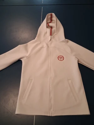 Vit hoodie med röd logga och dragkedja - Snygg vit hoodie från 2XU med hel dragkedja och två fickor framtill. Huva med röd rand på insidan och en liten röd logga på bröstet. Perfekt för en sportig och clean look.