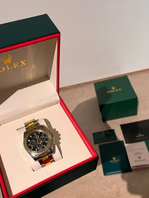 Role Daytona tvåtonad klocka - Snygg Role Daytona med tvåtonad länk i silver och guld. Klockan har svart urtavla med flera små ur och detaljer i guld. Kommer i originalbox med tillbehör och manualer. Perfekt för dig som vill ha en lyxig och klassisk accessoar.