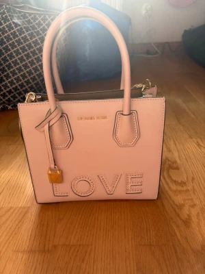 Ljusrosa Michael Kors handväska LOVE - Superfin ljusrosa handväska från Michael Kors med texten LOVE i nitar på framsidan. Väskan har både handtag och en lång axelrem, samt guldfärgade detaljer och ett litet hänglås. Perfekt för dig som vill ha en söt och trendig accessoar.
