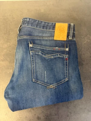 Blå raka jeans från Replay stl 34/30 - Säljer ett par klassiska blå jeans från Replay med raka ben och snygga slitningar. Jeansen har fem fickor, läderpatch bak i midjan och detaljerade sömmar på bakfickan. Tillverkade i bomull med en cool tvättad look.