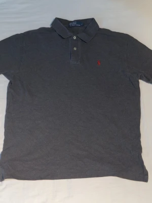 Mörkgrå pikétröja från Ralph Lauren - Klassisk mörkgrå pikétröja från Ralph Lauren med krage, två knappar och den ikoniska röda logotypen broderad på bröstet. Tillverkad i mjuk bomull för en bekväm känsla och perfekt passform. Enkel och stilren design som funkar till det mesta.