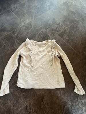 Ljusbeige långärmad topp med volang - Ljusbeige långärmad topp med volangdetaljer framtill. Tillverkad i mjukt bomullsmaterial och har en enkel, rund halsringning. Perfekt för dig som gillar en söt och stilren look. Plus frakt 