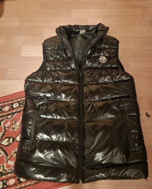 Svart dunväst från Moncler - Snygg svart dunväst från Moncler med glansig finish och klassisk logga på bröstet. Västen har dragkedja framtill, två sidofickor med dragkedja och en hög krage. Perfekt för lager-på-lager och riktigt trendig look. Tight fit