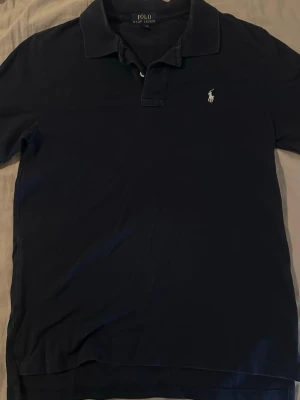 Marinblå pikétröja från Polo Ralph Lauren - Klassisk marinblå pikétröja från Polo Ralph Lauren med vit broderad logga på bröstet. Tröjan har krage, knappar vid halsen och korta ärmar. Perfekt till en avslappnad men ändå stilren look.