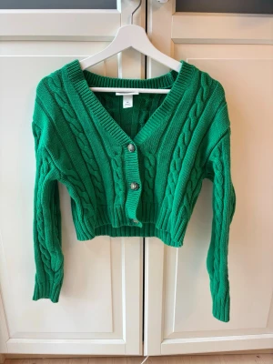 Grön kabelstickad kofta med knappar - Snygg grön kabelstickad kofta med tre stora knappar framtill. Koftan har v-ringning, ribbade muddar och är croppad i modellen. Perfekt för att lägga till lite färg i din outfit.