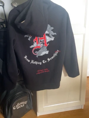 Svart hoodie från Hoodrich med tryck - Svart hoodie från Hoodrich med stora tryck på både fram- och baksida. Texten 'From Nothing To Something' och 'Product of the Streets' samt motiv av moln och änglar. Klassisk huva och känguruficka framtill. Perfekt streetwear-stil. Ny pris 1200