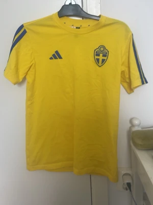 Svensk landslagströja Adidas gul - Gul svensk landslagströja från Adidas med blå detaljer och tryckt landslagsemblem på bröstet. Klassisk rund hals och korta ärmar, tillverkad i lätt och ventilerande material. Perfekt för fotbollsträning eller att bära på matchdag.