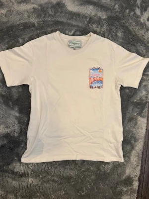 Vit t-shirt från Casablanca storlek S - Vit t-shirt från Casablanca med färgglad grafisk print på bröstet och ryggen som föreställer en pool och sol. T-shirten har rund hals och normal passform. Storlek S